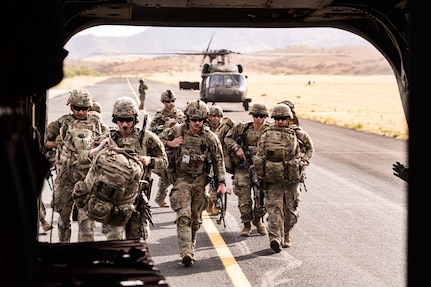 Multiple people wearing camouflage uniforms walk toward the rear of a helicopter while carrying duffle bags and rifles.