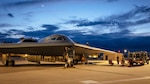Two airmen standing next to a truck on a runway refuel a B-2 aircraft with a crescent moon in a cloudy blue sky in the background.
