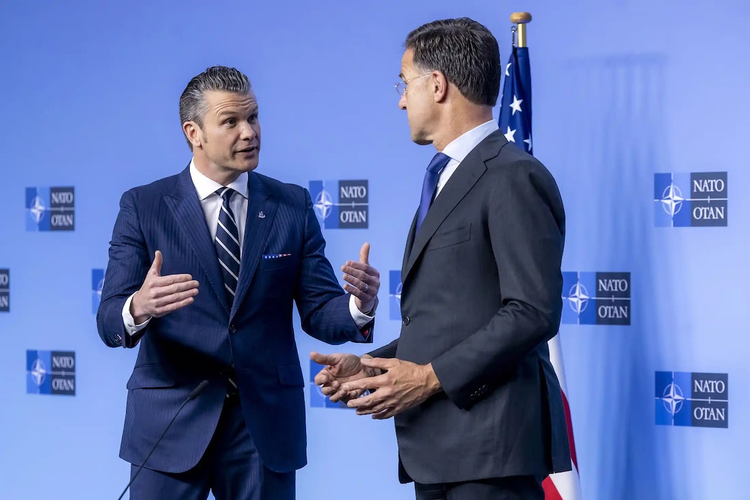Defense Secretary Pete Hegseth and another civilian stand and speak in front of a light blue wall with NATO logos.