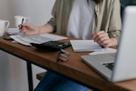Woman sits at desk in front of papers, notepad, calculator, glasses, and laptop.