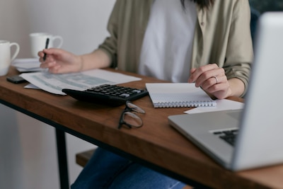 Woman sits at desk in front of papers, notepad, calculator, glasses, and laptop.