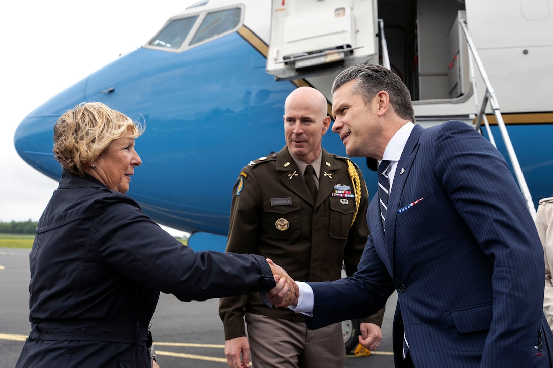 A man in a suit shakes hands with a woman outside an airplane while a man in military uniform watches during a cloudy day.