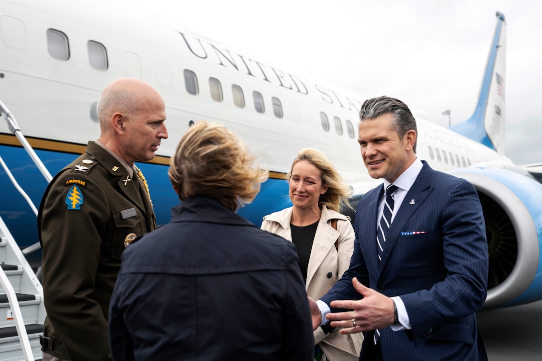 A man in a suit greets two people outside of an airplane with a woman standing behind during a cloudy day.