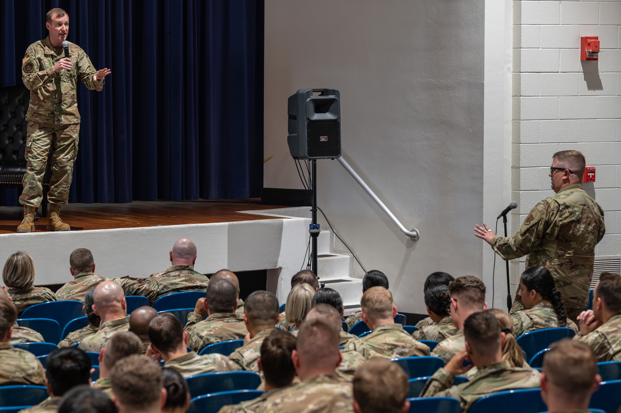 An individual in military uniform standing on a stage speaking to another individual in military uniform standing at a microphone.