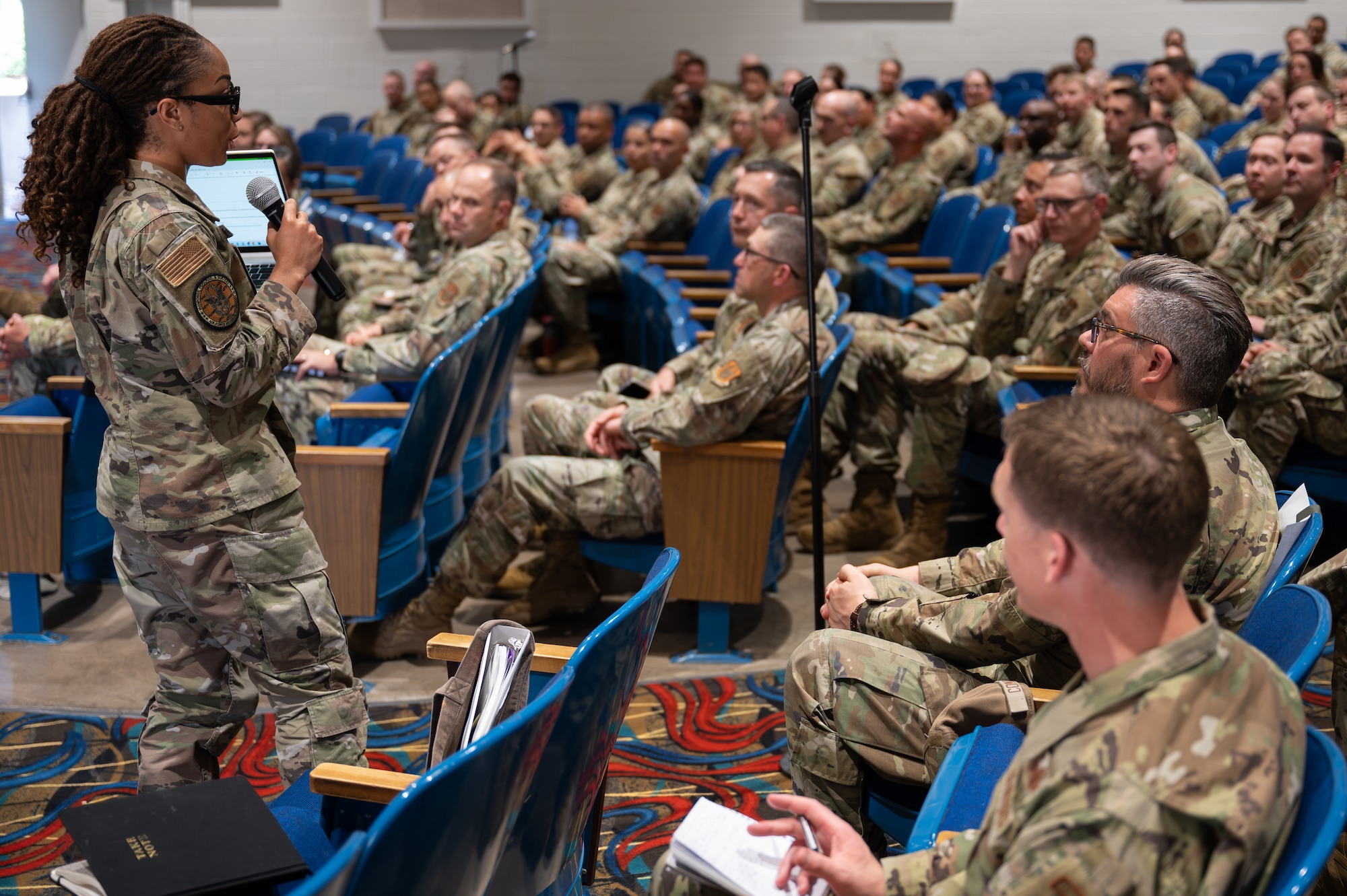 An individual in military uniform holding a microphone, speaks to an assembled crowd in an auditorium.