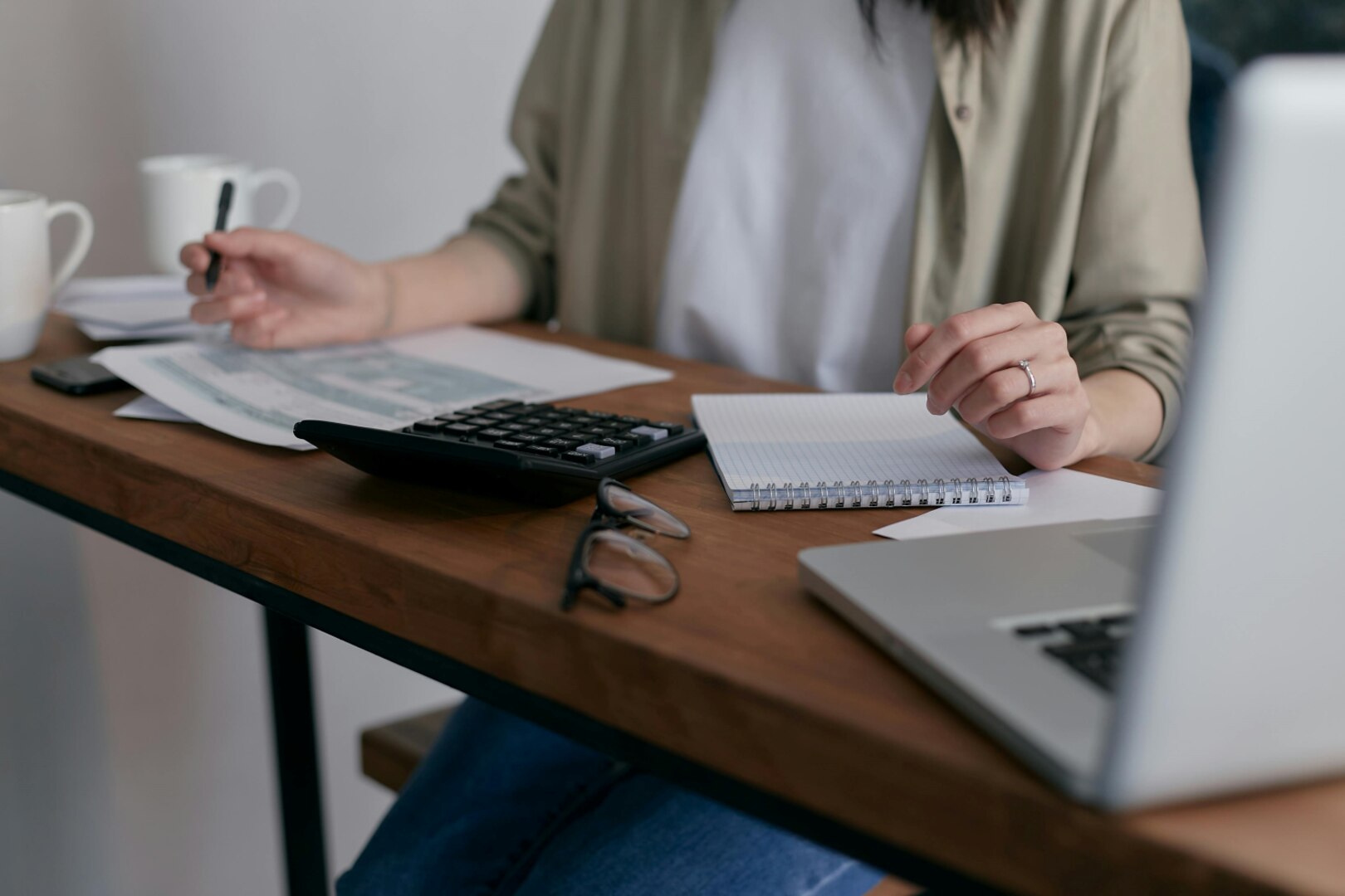 Woman sits at desk in front of papers, notepad, calculator, glasses, and laptop.