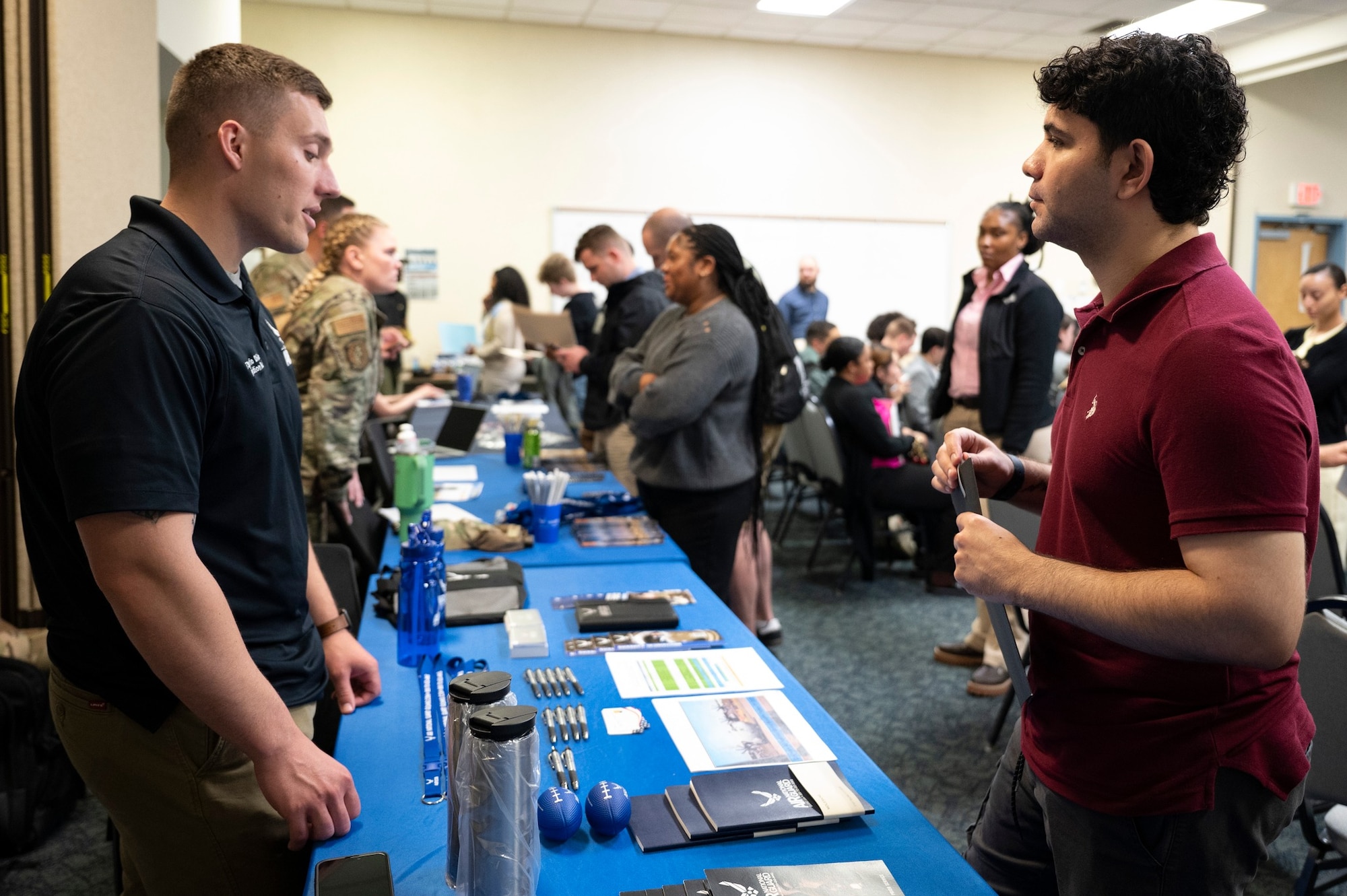 A man in a blue t-shirt on the left side of the photo speaks to a man in a maroon shirt on the right side of the photo with a table between them covered in pamphlets.