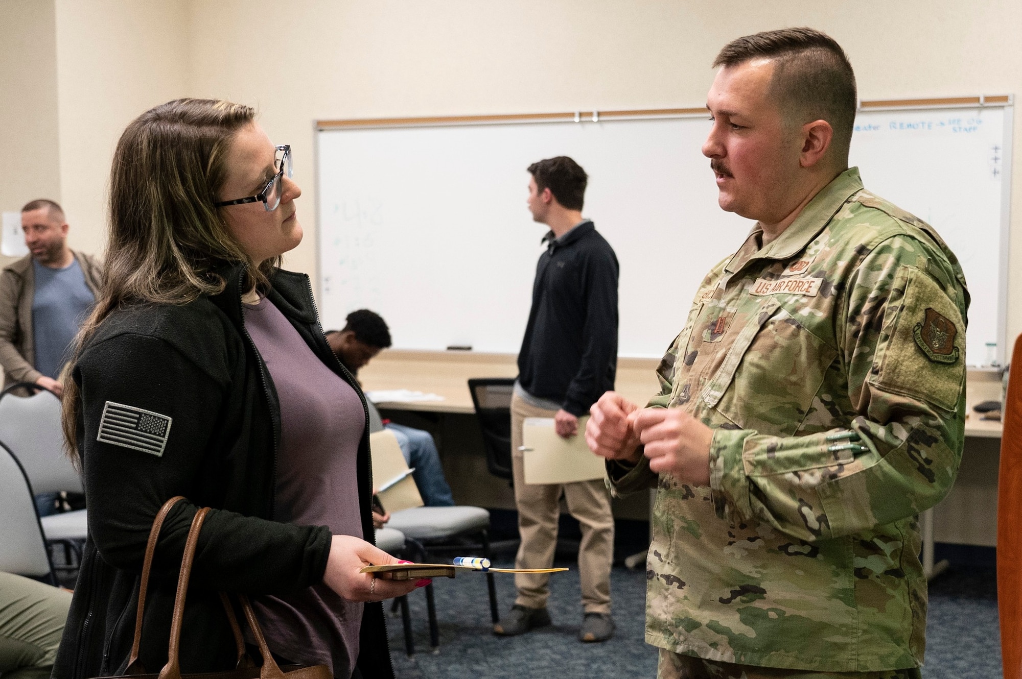 A man on the right side of the photo in an Air Force camouflage uniform speaks to a woman standing on the left side of the photo wearing a black sweater.
