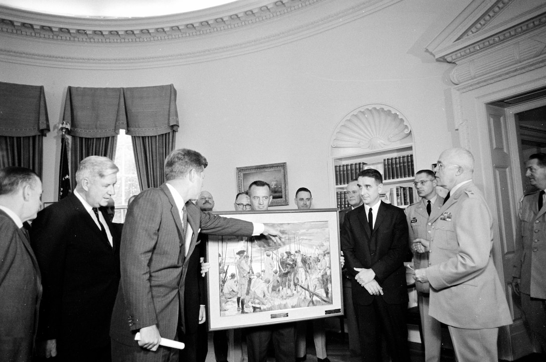 A dozen men stand in an oval office with a painting sitting on an easel in the center of their group.