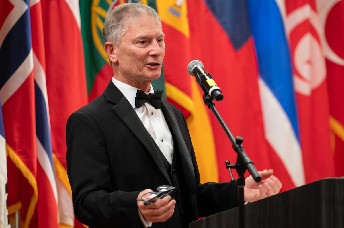 Man wearing tuxedo talks behind podium in front of many flags of various countries.