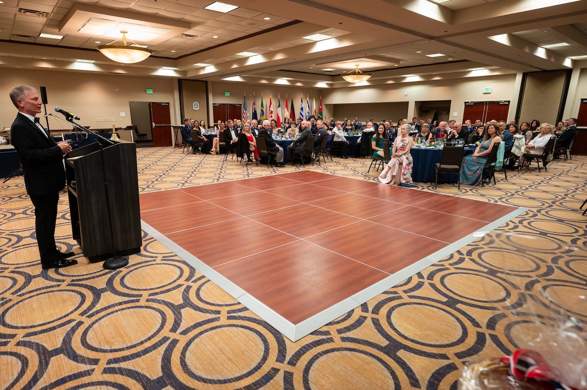 Man stands behind podium and addresses crowd sitting at round tables in ballroom.