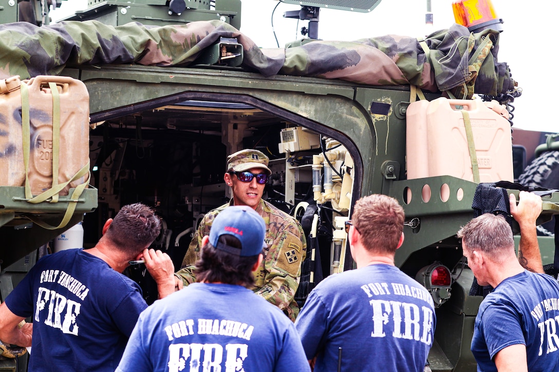 A soldier standing in front of a military vehicle talks to a group of firefighters wearing T-shirts outdoors.
