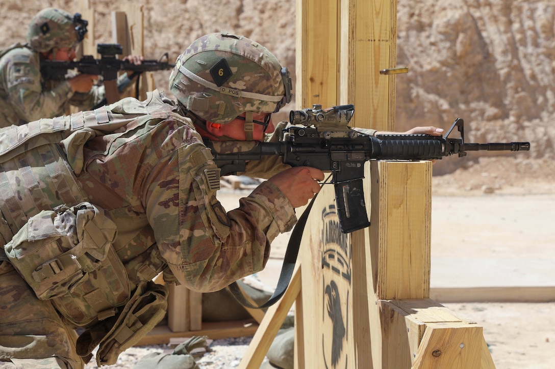 A soldier in tactical gear aims a military weapon on a wooden structure.
