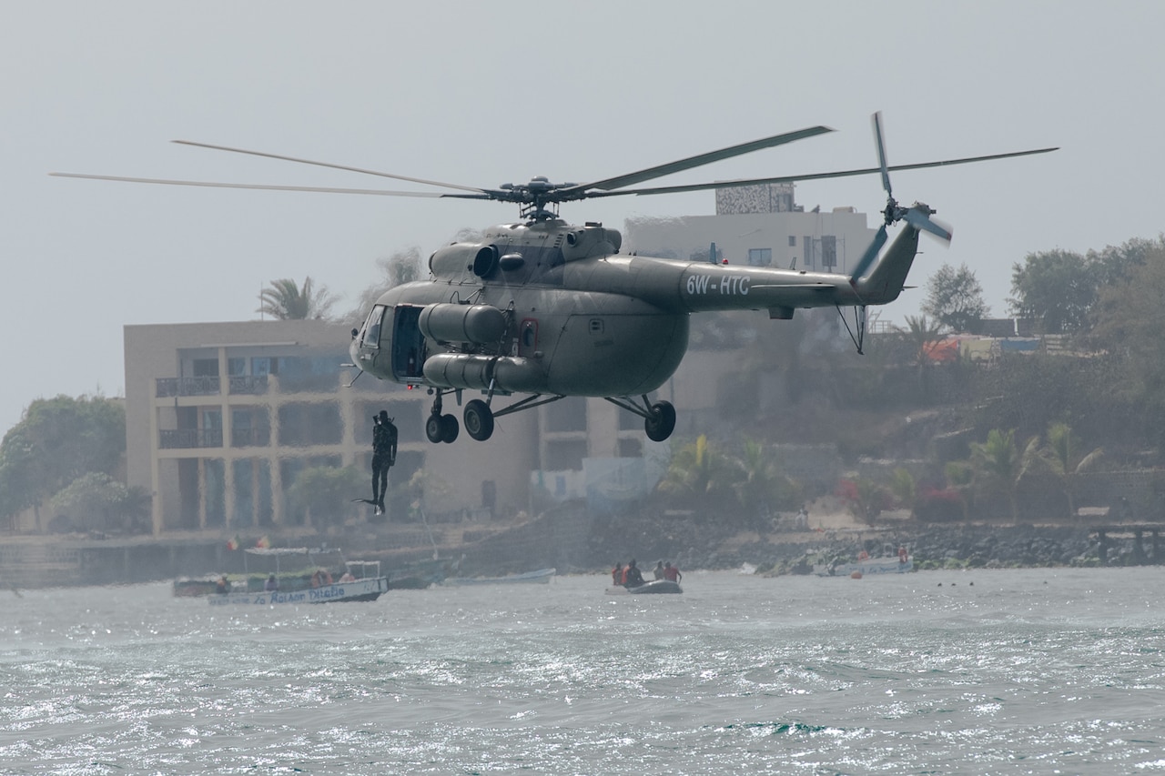 A sailor jumps into the ocean from a hovering helicopter. In the background are onlookers in boats and a coastline with hills, trees and buildings.