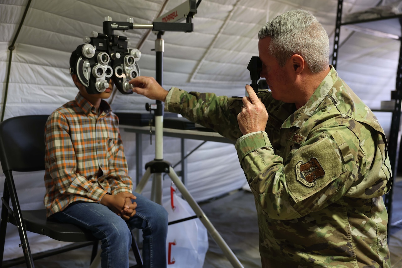 A man in uniform checks a boy’s eyes with a piece of equipment that has nine lenses that overlap inside a tent.