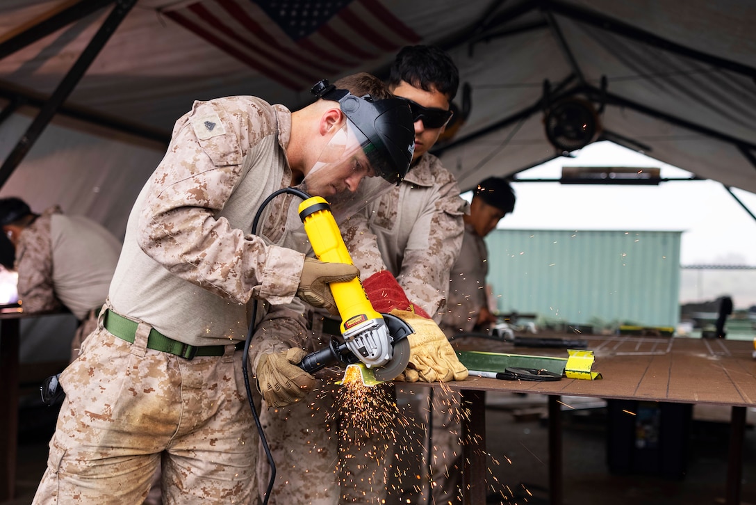 Sparks fly as a Marine uses a grinding tool on a metal stake as another person assists and others work in an outdoor tent.