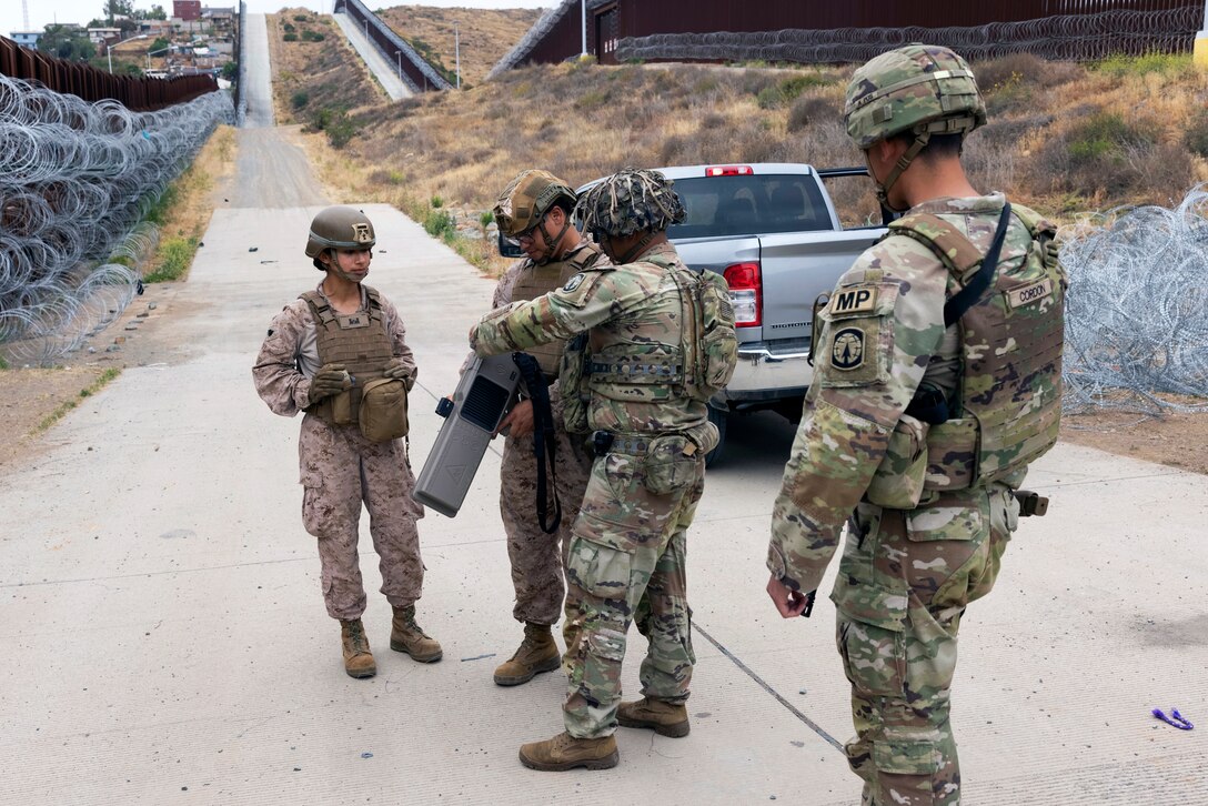 A soldier explains how to use an anti-drone device to two Marines as another soldier watches on a fence-lined road.