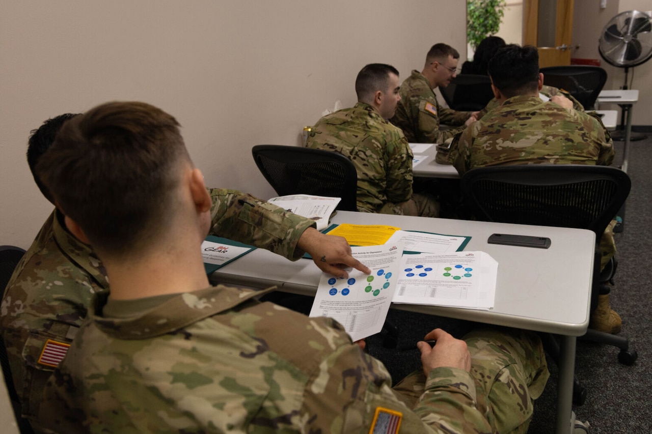 Soldiers in camouflage uniforms review course material while sitting at their desks in a classroom.