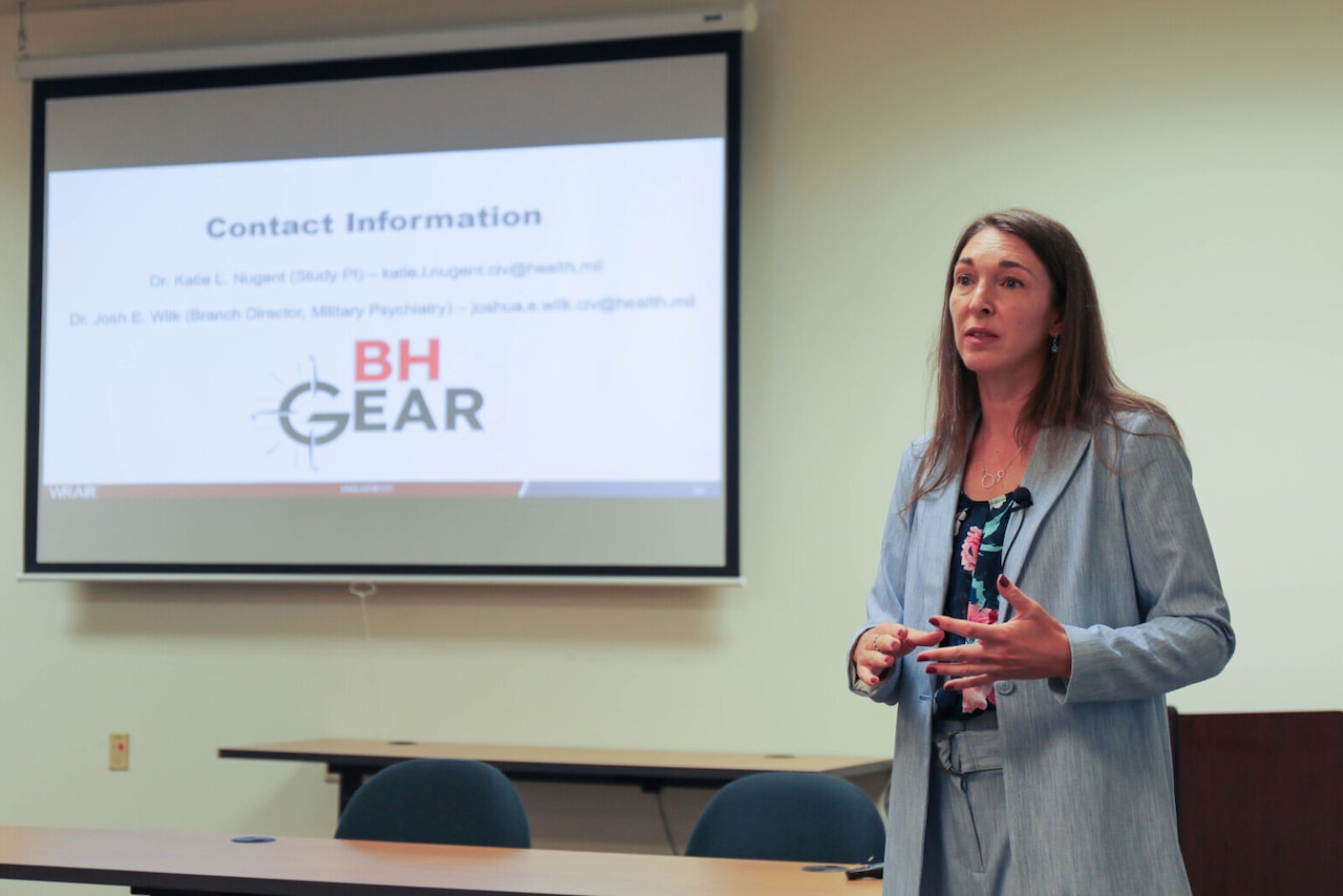 A woman in business attire speaks at the head of a classroom in front of a projector screen.