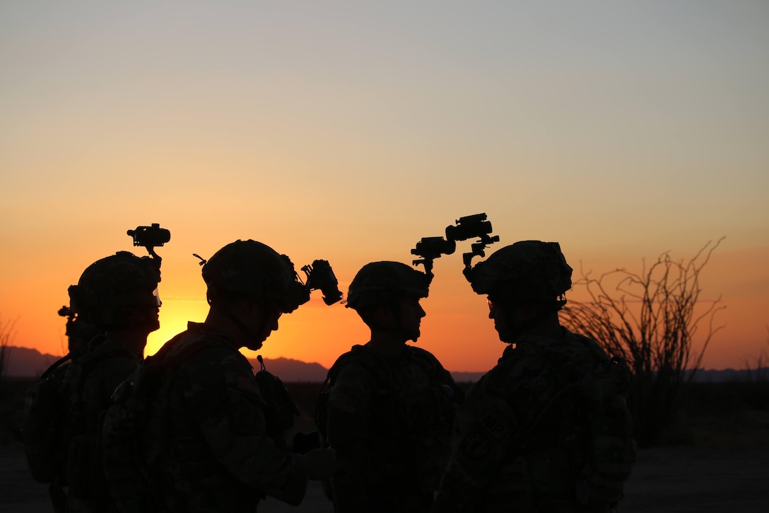 Four soldiers wearing helmets stand together, shown in silhouette against an orange sky.