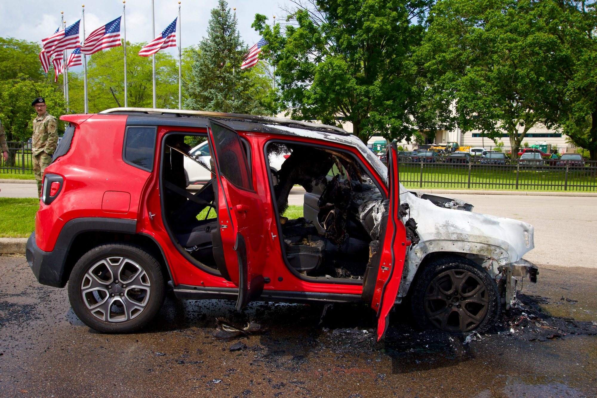 Red SUV with front half severely damaged by fire, showing melted and charred engine components. Both front doors are open. Two uniformed Security Forces members stand nearby in the background, observing the scene. American flags wave in the distance under a partly cloudy sky.