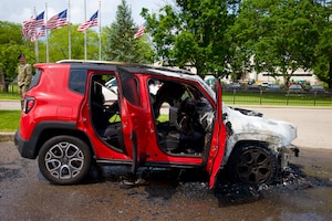 Red SUV with front half severely damaged by fire, showing melted and charred engine components. Both front doors are open. Two uniformed Security Forces members stand nearby in the background, observing the scene. American flags wave in the distance under a partly cloudy sky.