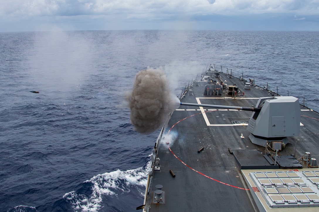 Smoke billows from the tip of a large gun mounted on the deck of a ship at sea as it fires a projectile on a cloudy day.