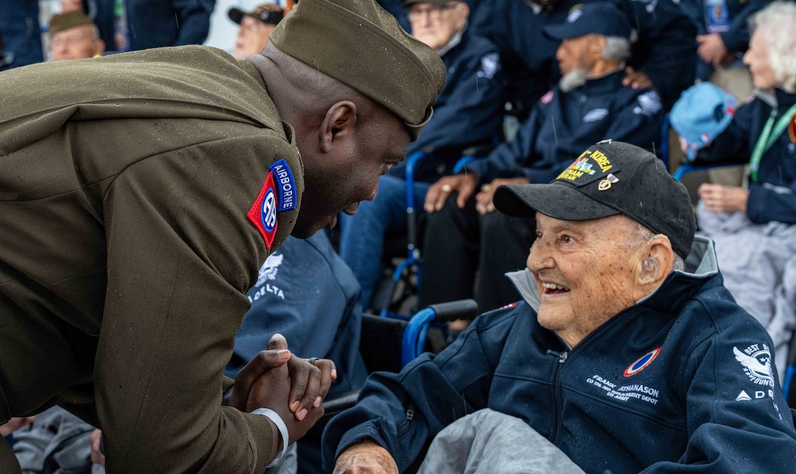 A soldier leans over to speak to a seated World War II veteran wearing a light jacket and hat as other veterans and people sit in the background.