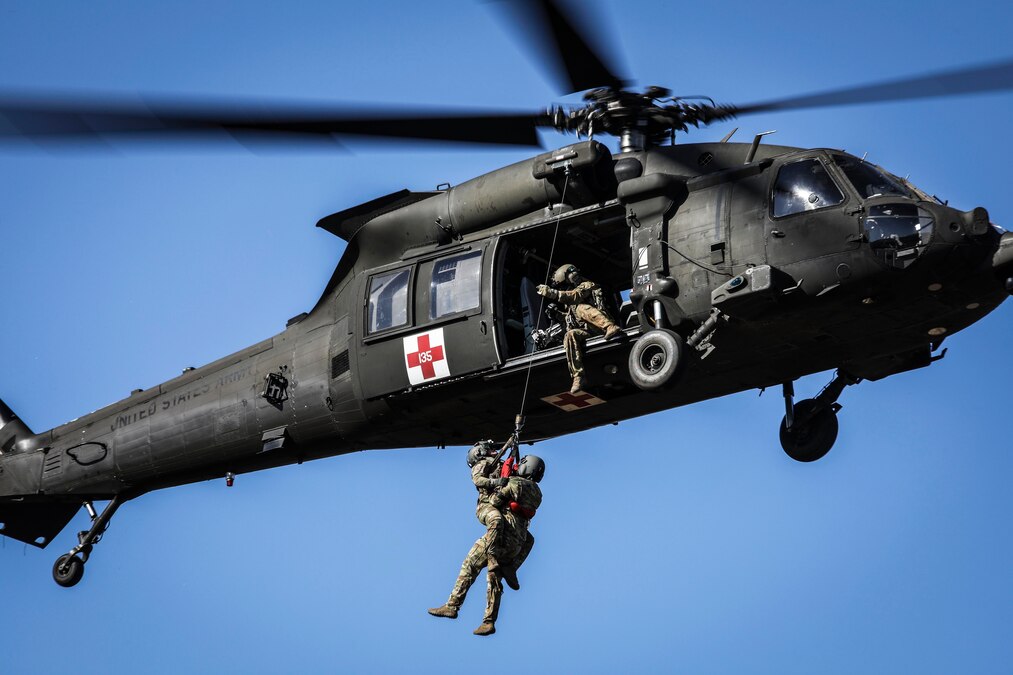 A soldier seated at an open door of a flying helicopter guides two others strapped to a hoist line up to the door on a cloudless day.