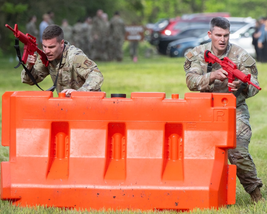 Two men brandishing red, fake guns take cover behind an orange barricade.
