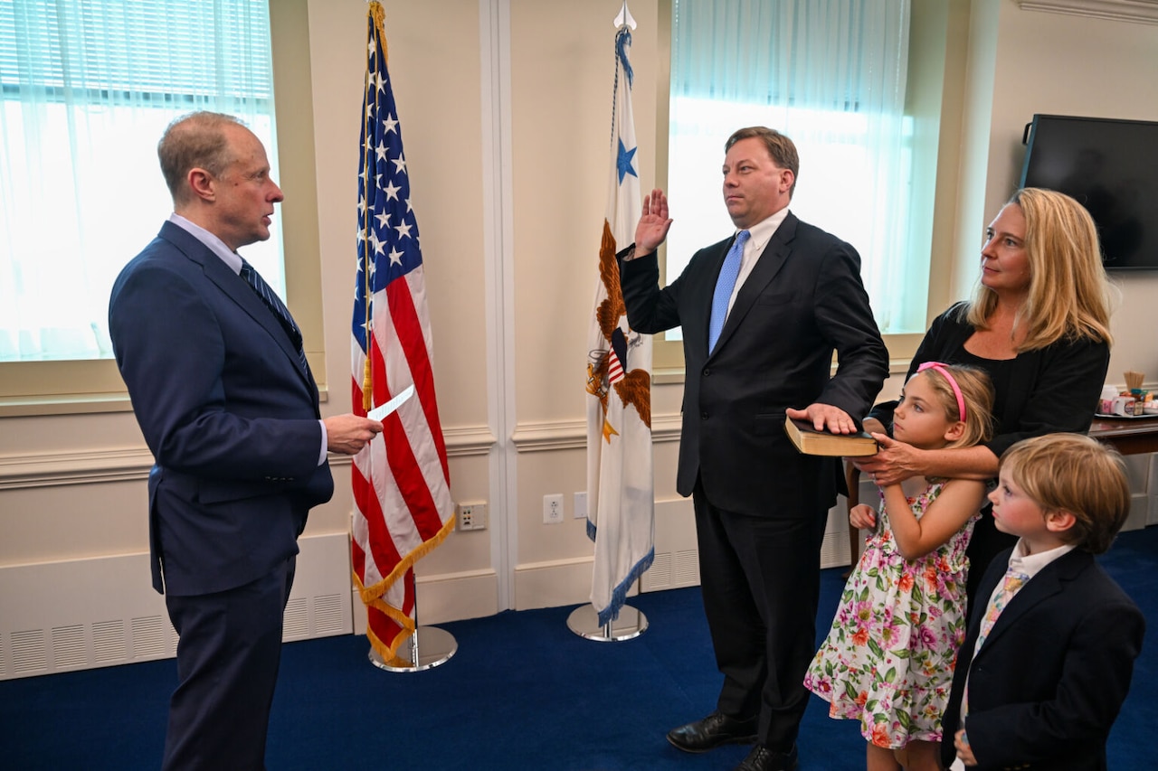 A man in a suit takes an oath, raising his right hand, as another man in a suit administers the oath. They stand in front of U.S. and presidential flags. A woman, a young girl in a dress, and a young boy in a suit watch the indoor ceremony.