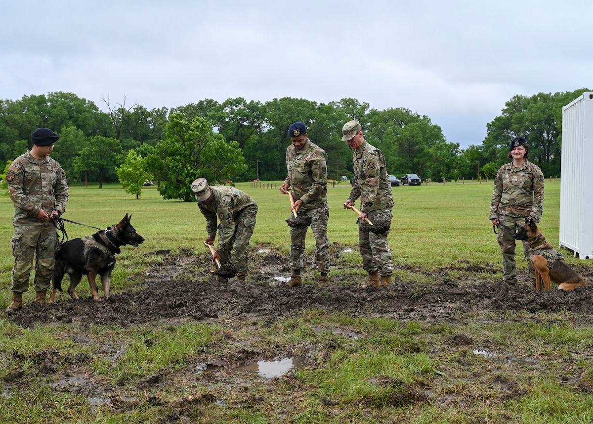 Men in military uniforms use golden shovels to move dirt while two people in military uniforms and their military working dogs stand next to them.