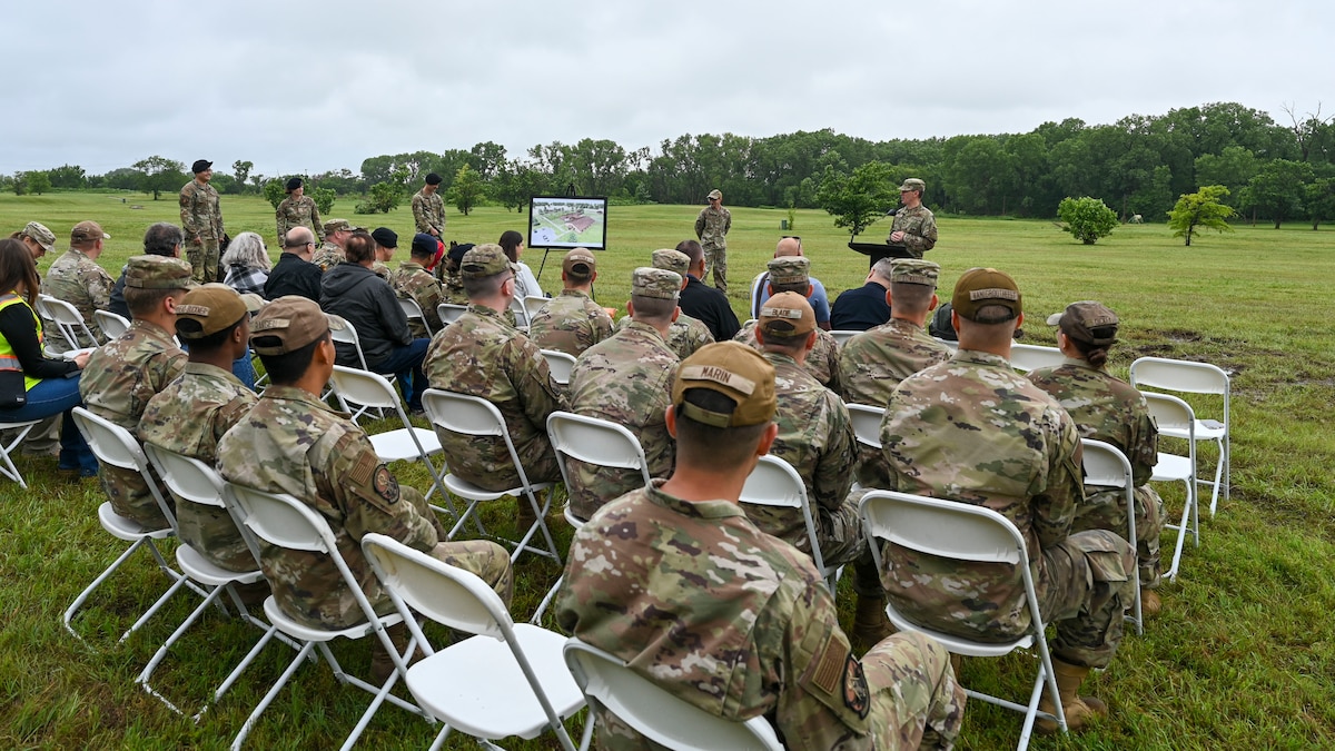 People in military uniforms and civilian clothes sit in chairs while a man in a military uniform speaks at a podium in front of them.