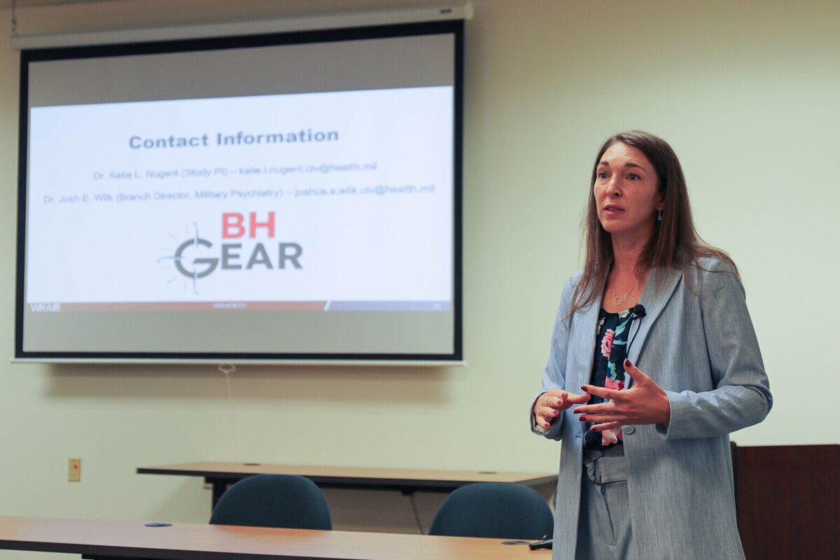 A woman in business attire speaks at the head of a classroom in front of a projector screen.
