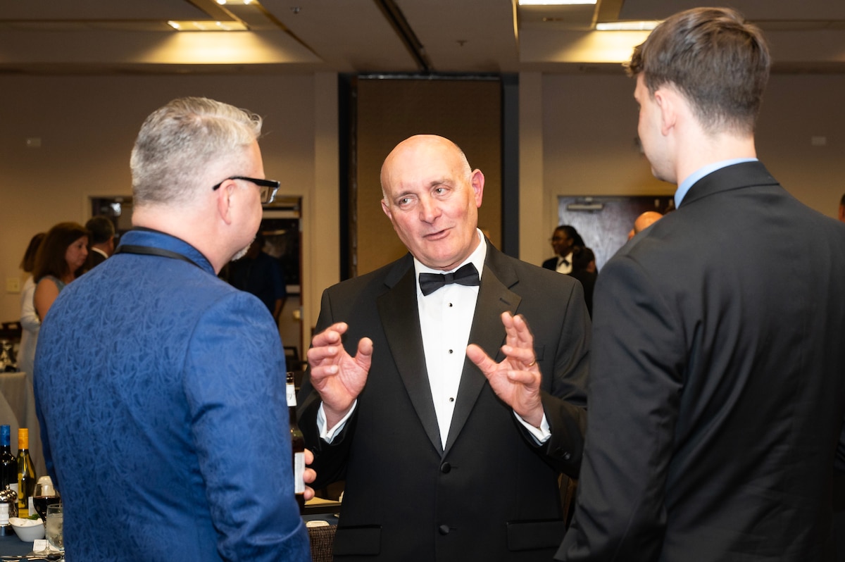 Three men standing, talking. Two have backs to camera. Man facing camera is wearing a tuxedo.