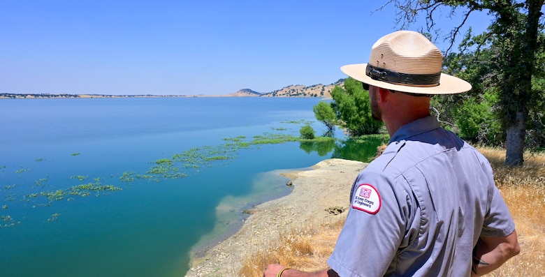 park ranger in foreground looks out toward blue lake from the shore.