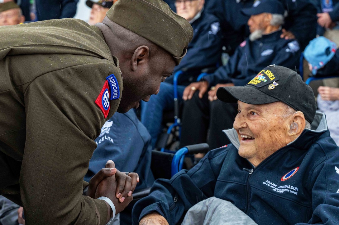 A soldier leans over to speak to a seated World War II veteran wearing a light jacket and hat as other veterans and people sit in the background.