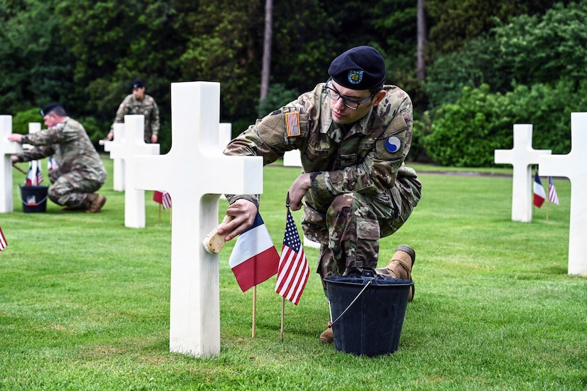 A soldier washes a tombstone shaped like a cross outside.