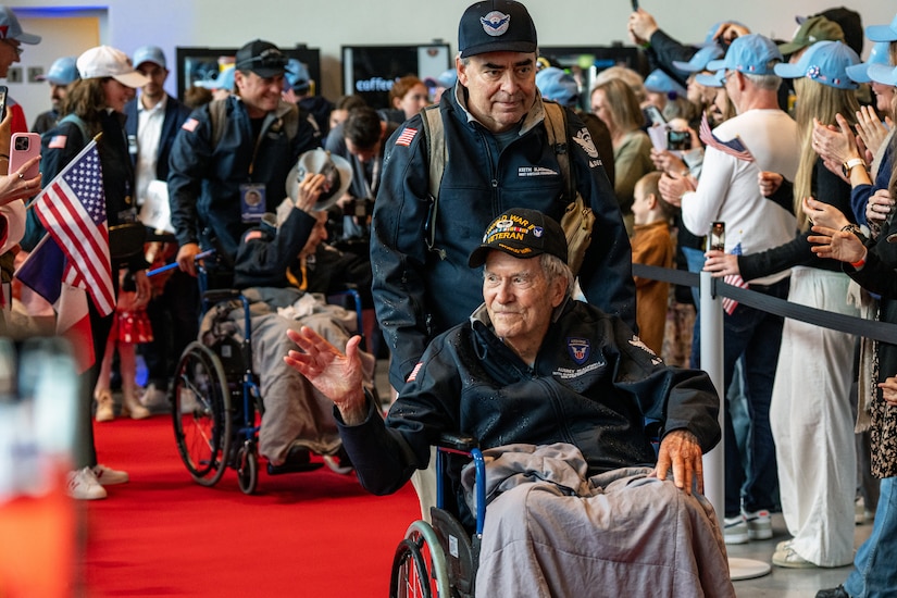 A veteran is wheeled in on a red carpet as a crowed cheers holding U.S. flags and phones.