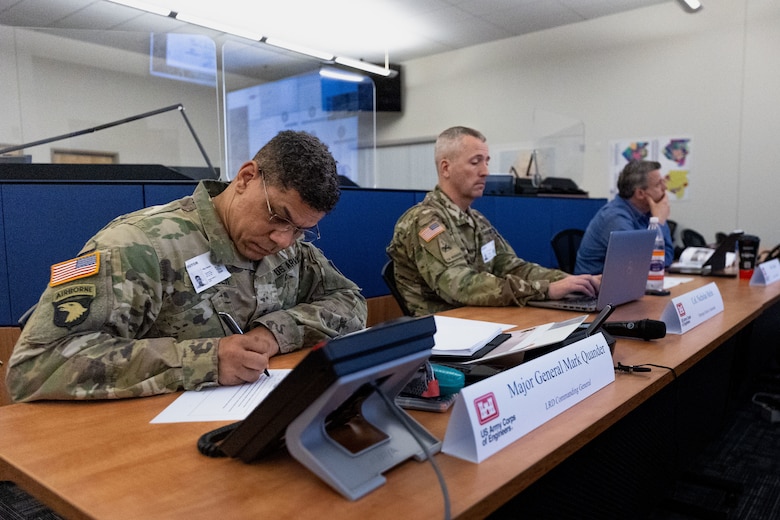 Three men sit at a table watching a presentation.