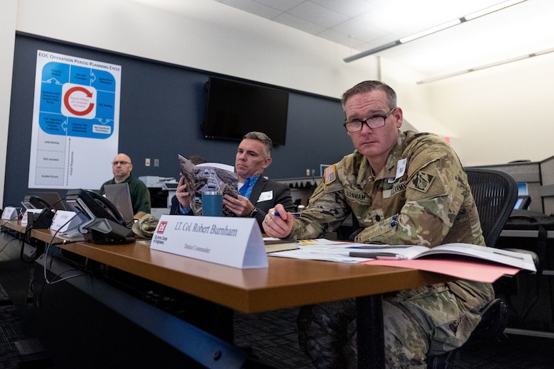 Three men sit at a table watching a presentation.