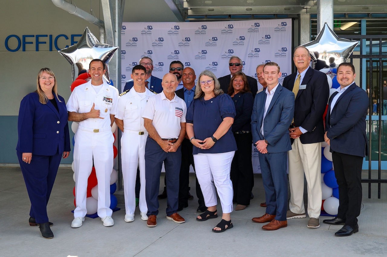 A group of adults, including two sailors in uniforms, district officials in business attire and a woman, stand together smiling in front of a school.