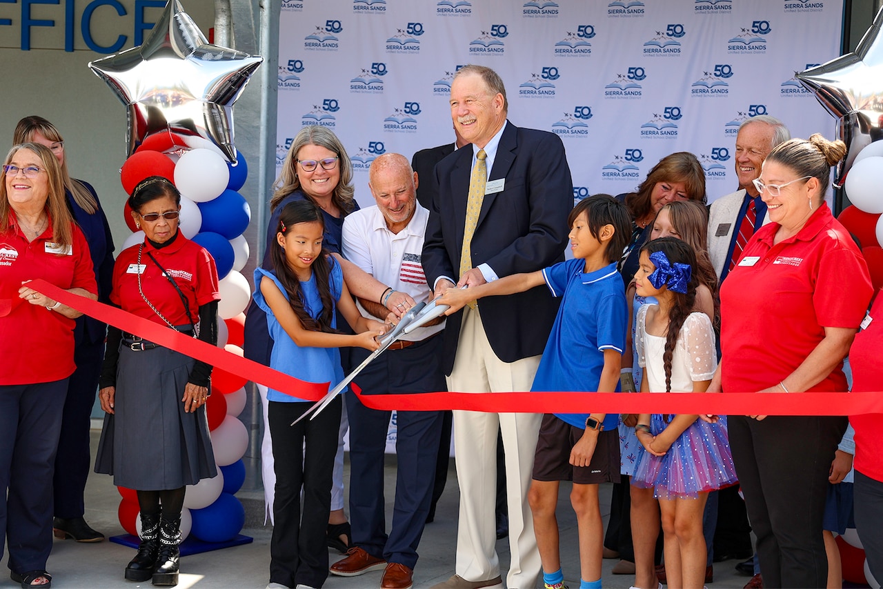 A group of people, including children in shirts and adults, stand together holding a large ribbon.
