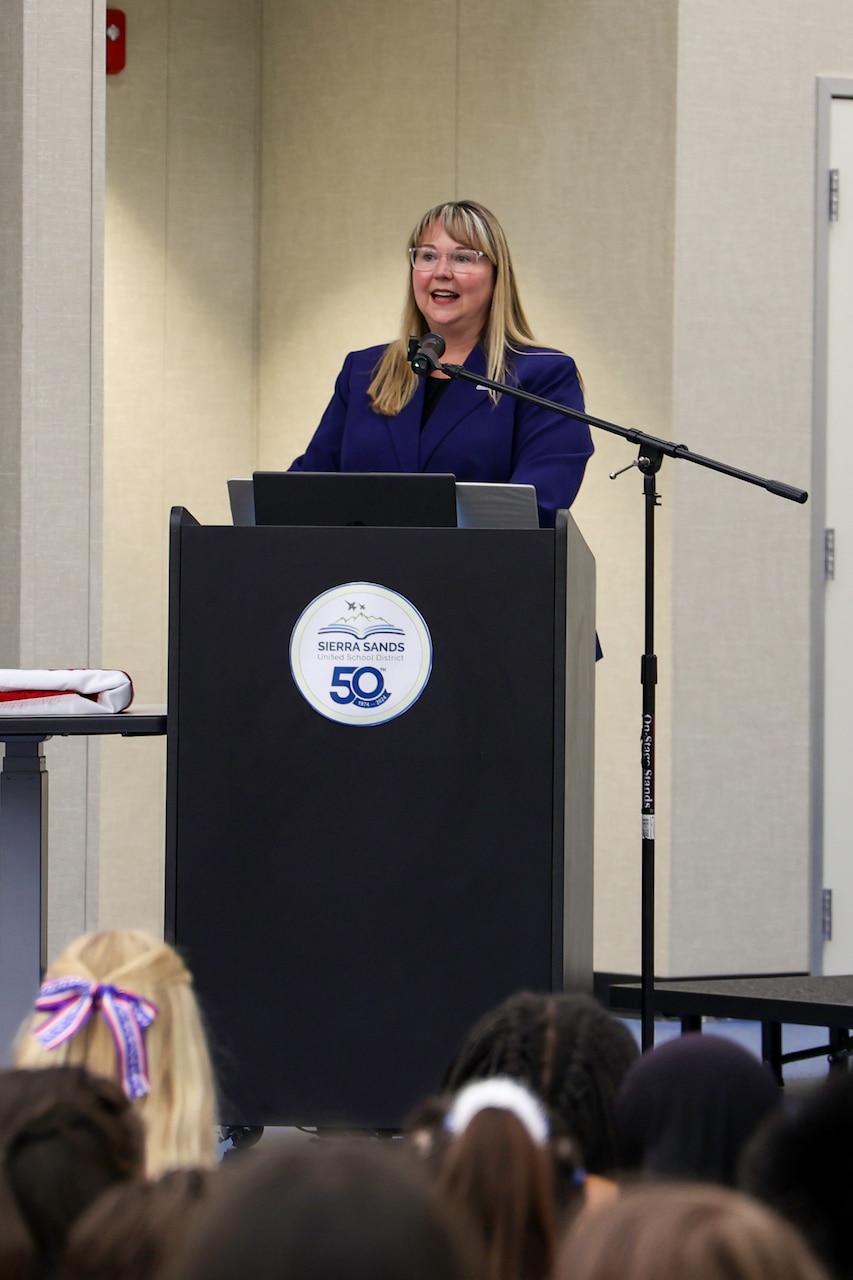 A woman in a blazer stands at a podium with a microphone, speaking to an audience.