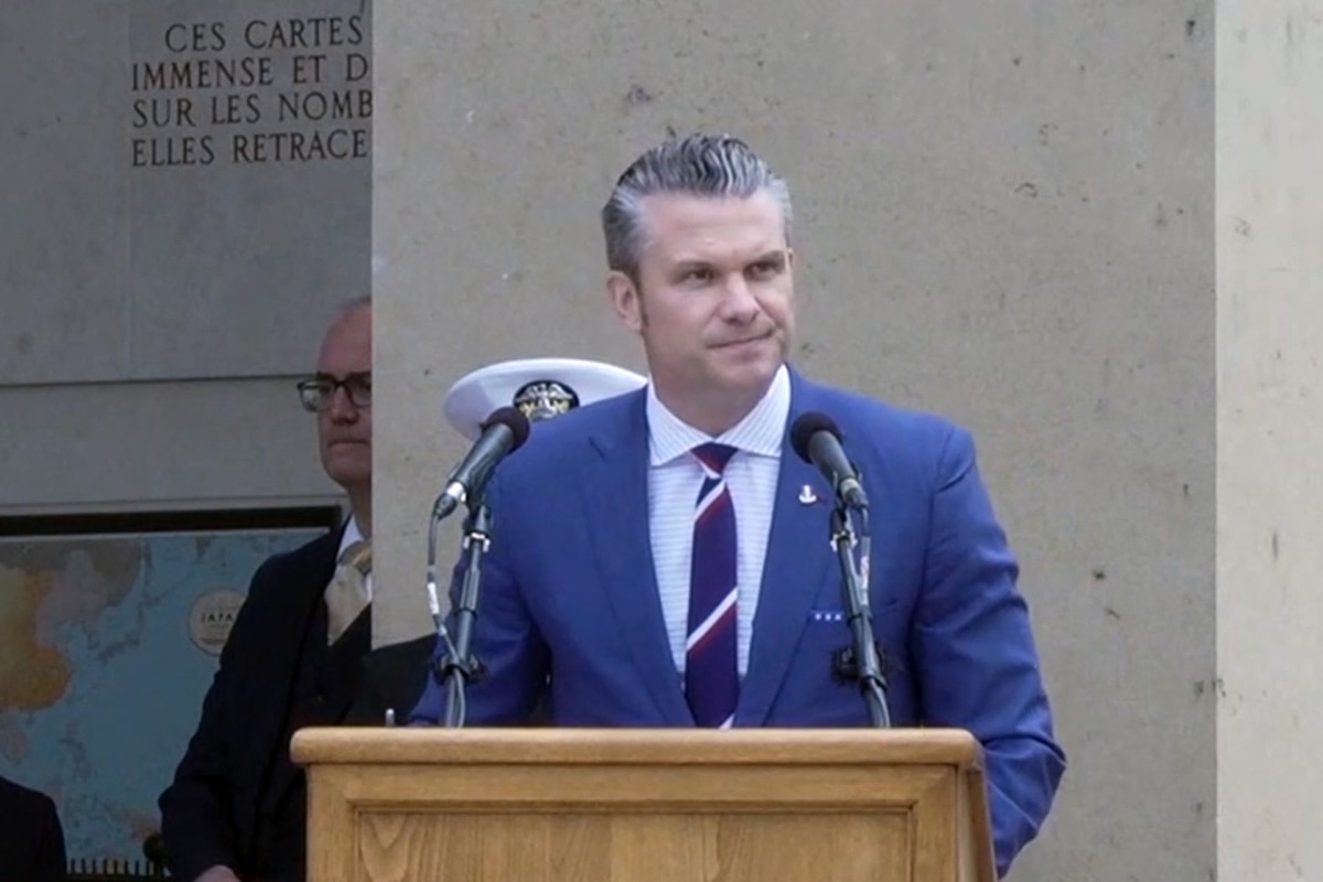 Defense Secretary Pete Hegseth stands at a lectern with two attached microphones outside a building.