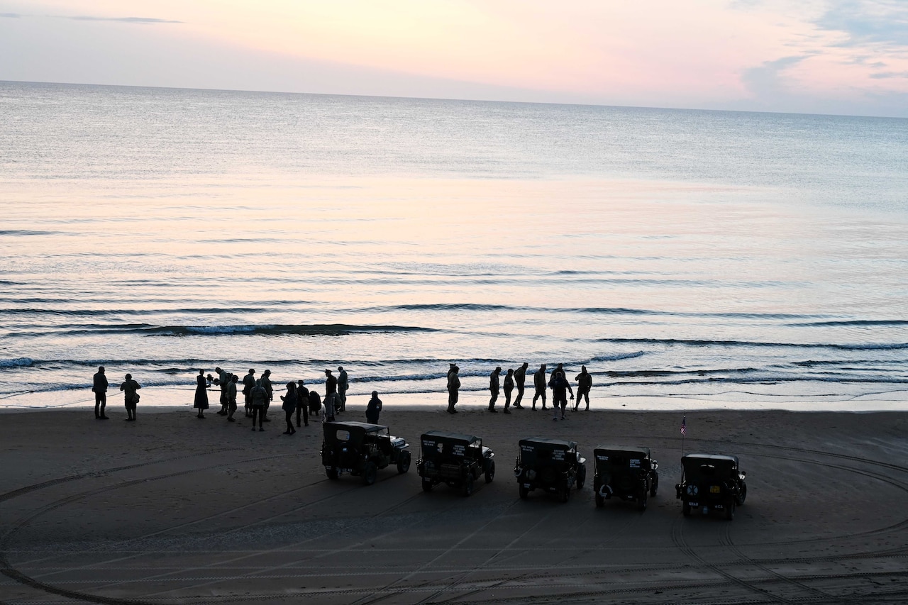 Historical vehicles and people in World War II outfits line a beach during lowlight.