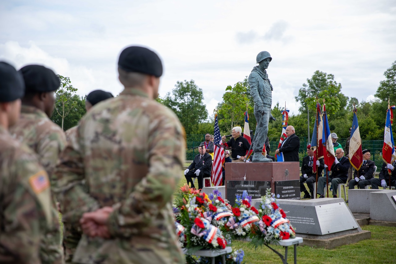 Soldiers stand in formation in front of a monument of a soldier wearing a helmet.