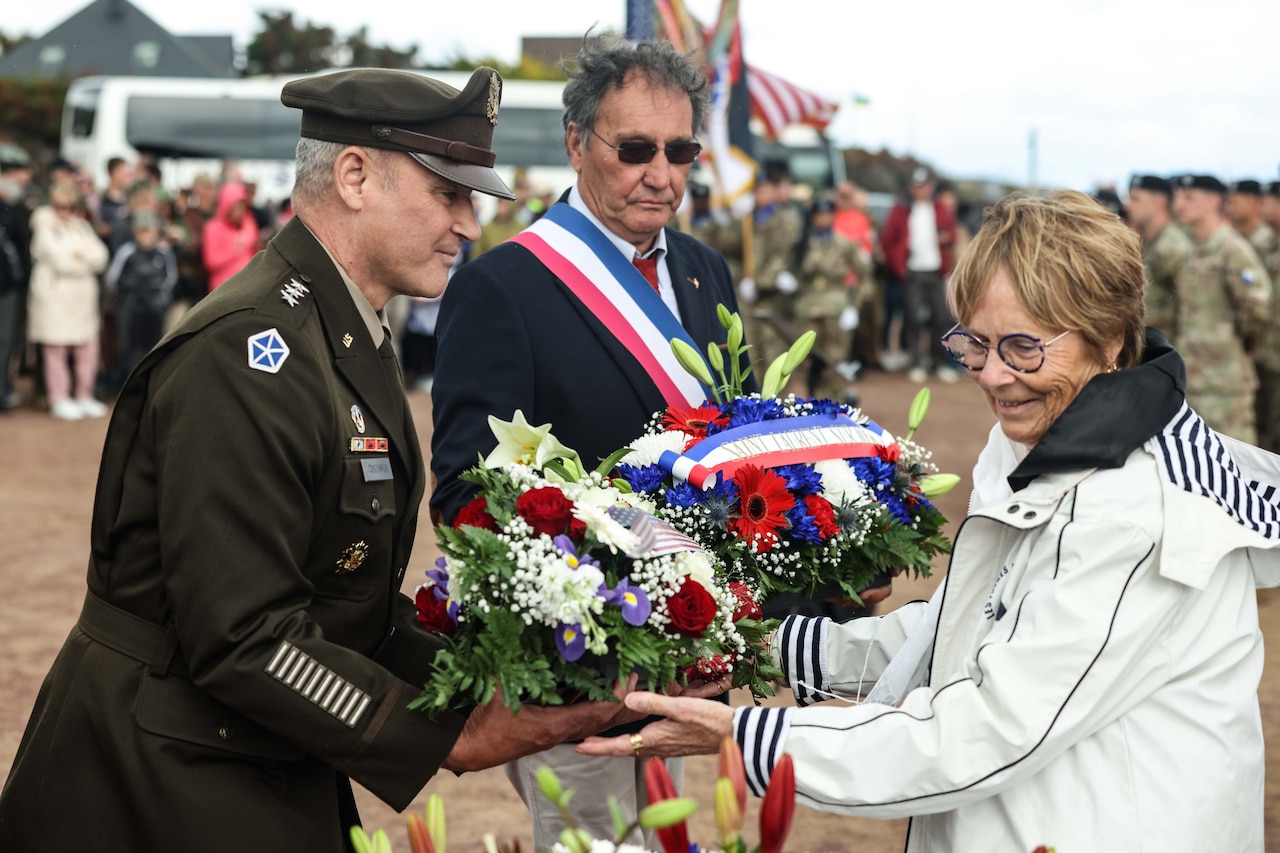 A general helps a woman carry a wreath outside while a man wearing a suit and a blue, white and red sash looks on.