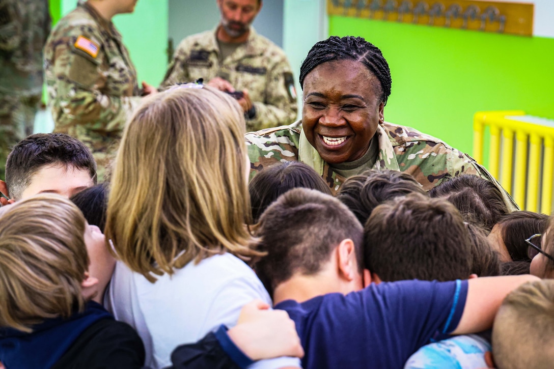 A soldier smiles and hugs a group of children in a classroom as other soldiers stand in the background.