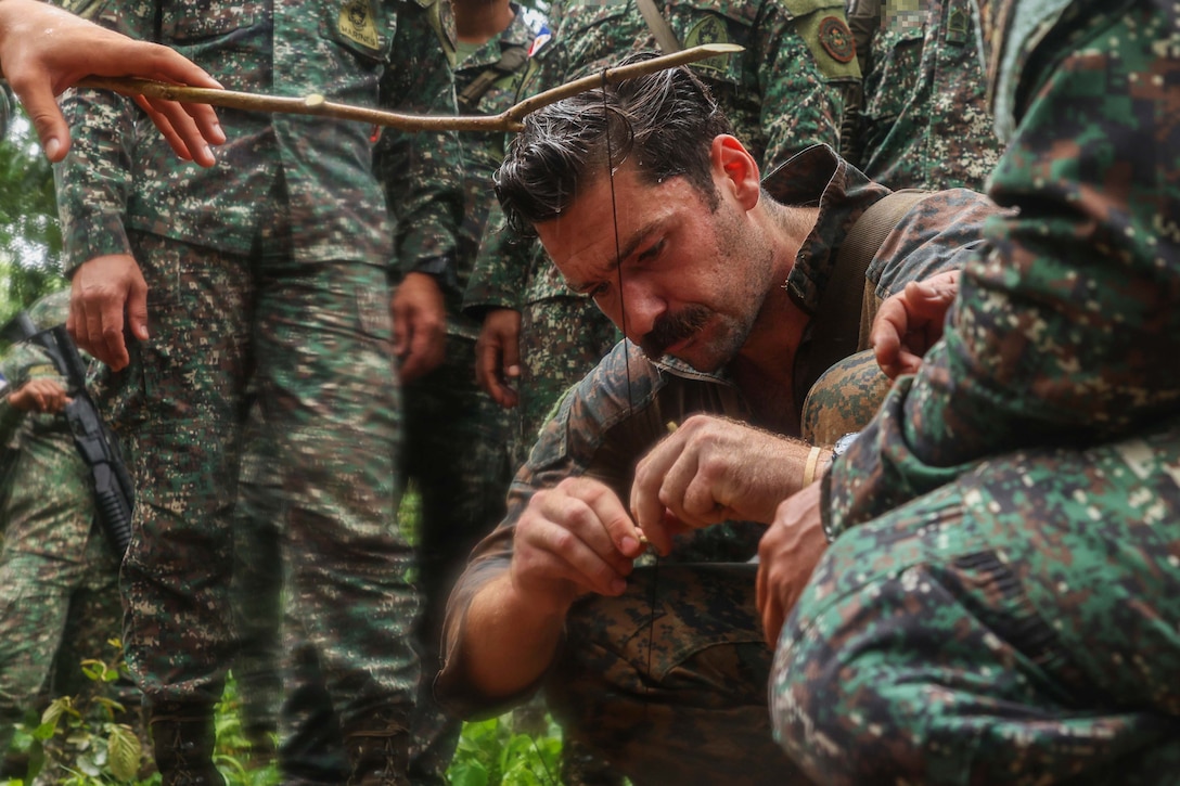 A sailor sets a trip wire hanging from a stick in a jungle setting as other troops watch in the background.